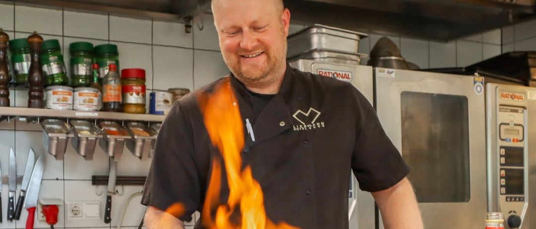 Ein Koch kocht in einer Küche mit Flammen aus einer Bratpfanne, lächelt, während verschiedene Gewürze und Küchenutensilien an der Wand hängen.