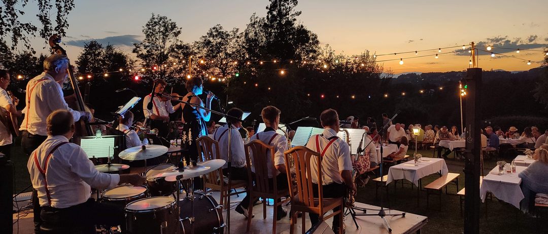 Ein Abendband spielt draußen mit Schlagzeug, Violinen und Saxophonen. Gäste sitzen um Tische unter einem Blätterdach mit Lichterketten.