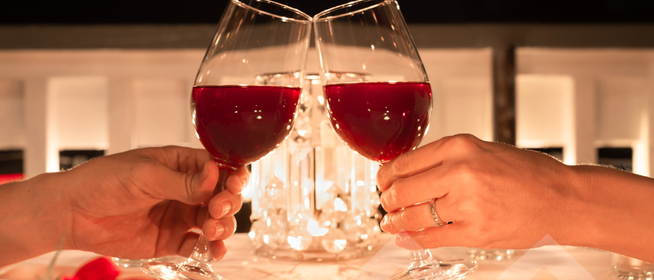 Zwei Hände stoßen mit Gläsern roten Wein an, neben einer Kerze und einer Rose. Die Umgebung ist intim, mit gedämpfter Beleuchtung und einer romantischen Atmosphäre.