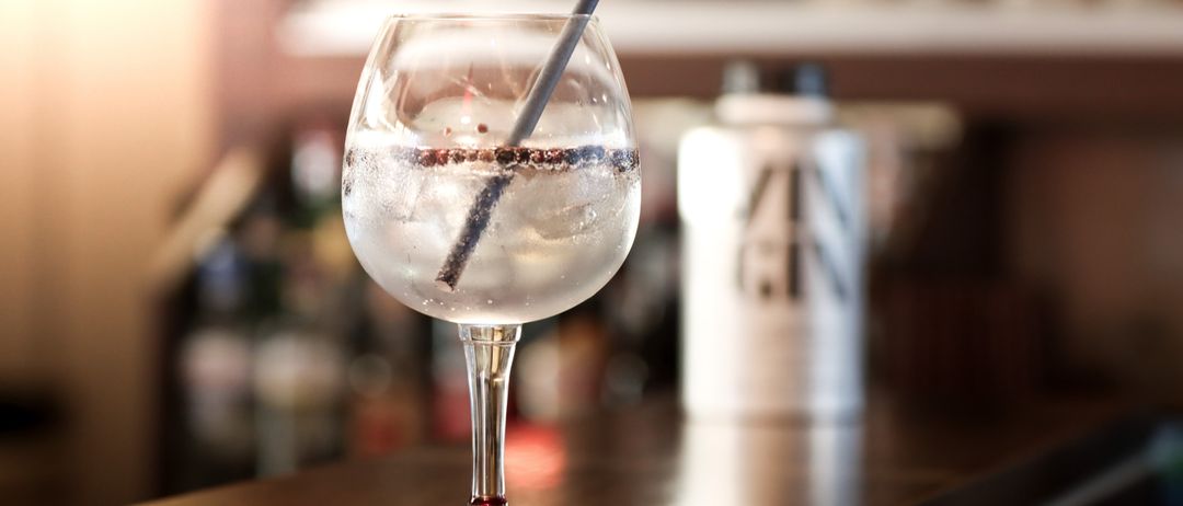 Ein Weinglas mit einem Cocktail, garniert mit Eis und einem Strohhalm, auf einer hölzernen Bar. Dahinter ist eine verschwommene Flasche mit dem Etikett 'GIN'.