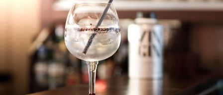 Ein Weinglas mit einem Cocktail, garniert mit Eis und einem Strohhalm, auf einer hölzernen Bar. Dahinter ist eine verschwommene Flasche mit dem Etikett 'GIN'.