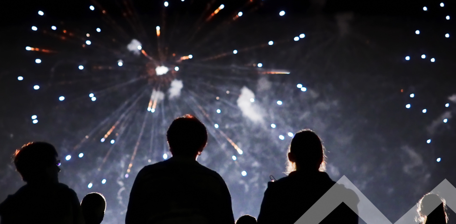 Eine Gruppe von Menschen beobachtet ein Feuerwerk bei Nacht. Die Feuerwerke explodieren am Himmel und beleuchten die Menge unten.