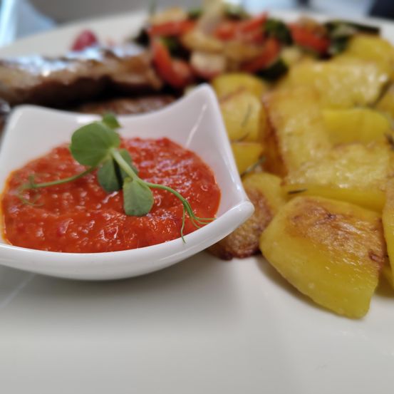 Auf einem weißen Teller befinden sich gegrilltes Fleisch, geröstete Kartoffeln und eine rote Sauce mit grüner Garnierung.