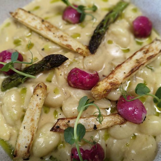 Ein Teller mit cremiger Pasta aus weißen Bohnen, garniert mit geröstetem Spargel und Radieschen. Das Gericht ist außerdem mit einigen kleinen grünen Blättern verziert.