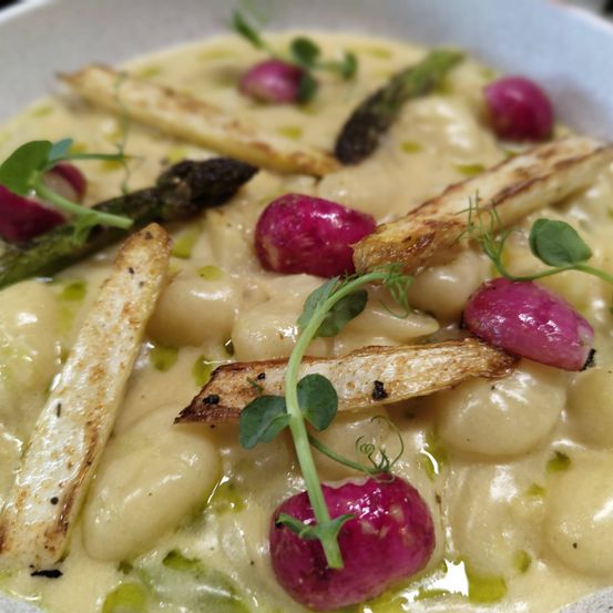 Eine Schüssel mit cremigen Gnocchi, gerösteten Radieschen, Spargel und Kräutern. Das Gericht ist mit grünen Blättern und einem Hauch von Öl garniert.