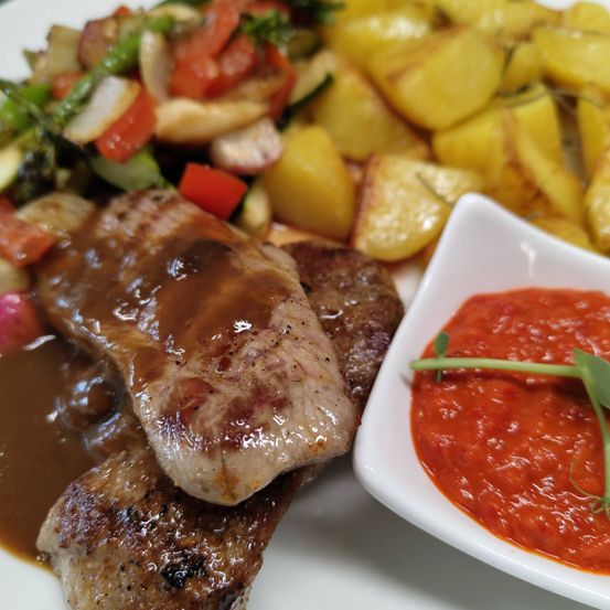 Ein Teller mit gegrilltem Fleisch in Sauce, begleitet von einer Beilage gebratener Kartoffeln und einem gemischten Gemüsesalat. Eine kleine Schüssel mit roter Sauce, verziert mit einem grünen Blatt.