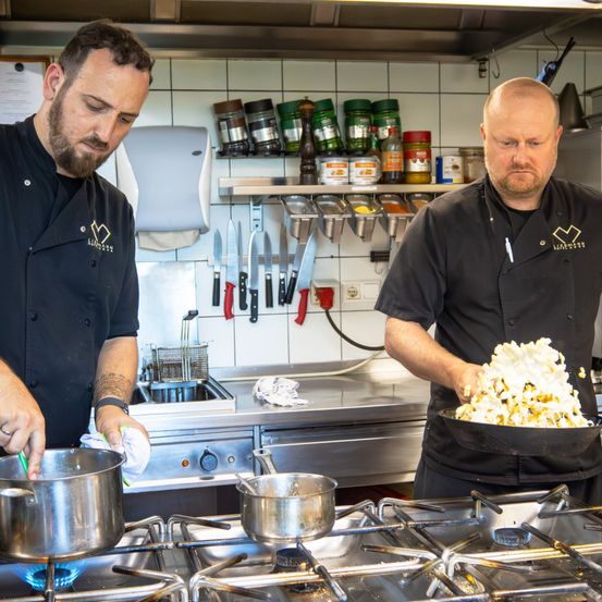 Zwei Köche in einer Küche, die Essen zubereiten, einer rührt einen Topf, der andere mischt Essen in einer Pfanne, umgeben von Küchenutensilien und Zutaten.