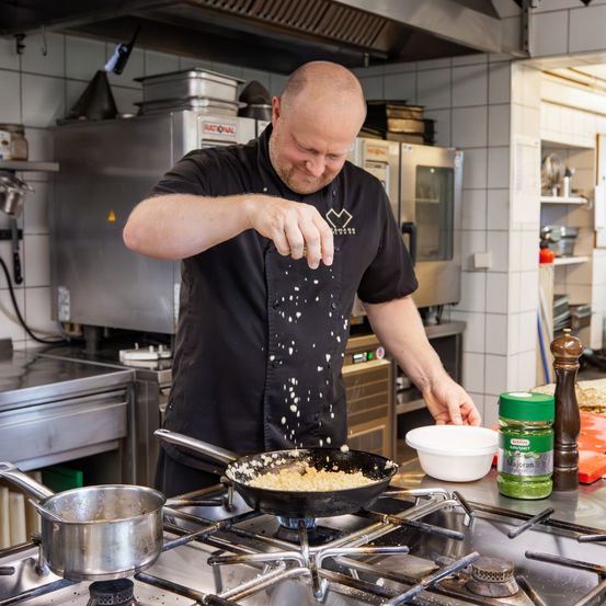 Ein Koch in einer schwarzen Uniform würzt Essen in einer Pfanne auf einem Gasherd in einer Küche.