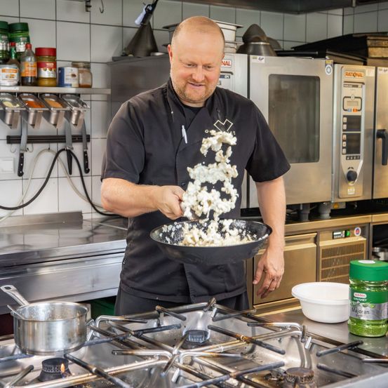 Ein Koch in einem schwarzen Hemd und einer Schürze bereitet in einer Küche mit Öfen, Töpfen und einem Herd Essen zu. Er hält eine Schüssel mit Zutaten.