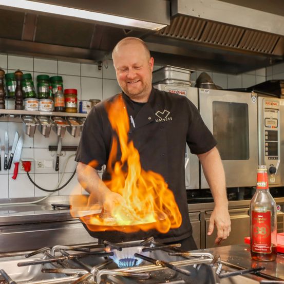 Ein Koch mit einem flammenden Wok in der Küche. Er lächelt und trägt ein schwarzes Kochuniform. Eine Flasche 38 Original Chili Beer steht auf dem Tresen.