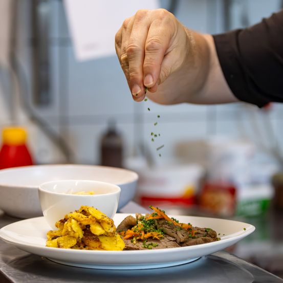 Ein Koch streut grüne Kräuter über einen Teller mit Kartoffeln und Fleisch. Ein Schüssel und eine Flasche sind im Hintergrund.