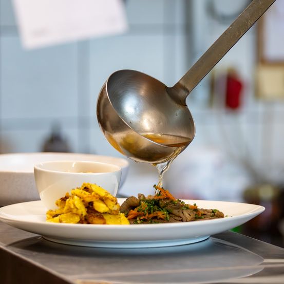 Ein Koch gießt Sauce aus einem Schöpflöffel über ein Gericht auf einem weißen Teller mit frittierten Kartoffeln und Gemüse. Ein Schüssel steht in der Nähe.