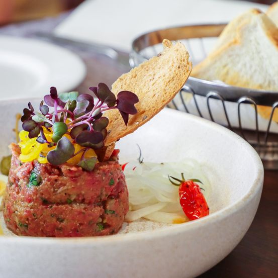 Eine Nahaufnahme eines Gerichts in einer Schüssel mit einer Garnitur aus Microgreens, einer Scheibe Brot und Zwiebeln. Das Gericht ist mit einer roten Tomate garniert.