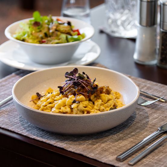 Eine Schüssel mit Käsepasta, mit Grünzeug garniert, steht auf einem Platzset mit Salat und Besteck. Eine weitere Schüssel mit Salat und ein Glas befinden sich auf einem Untersetzer.