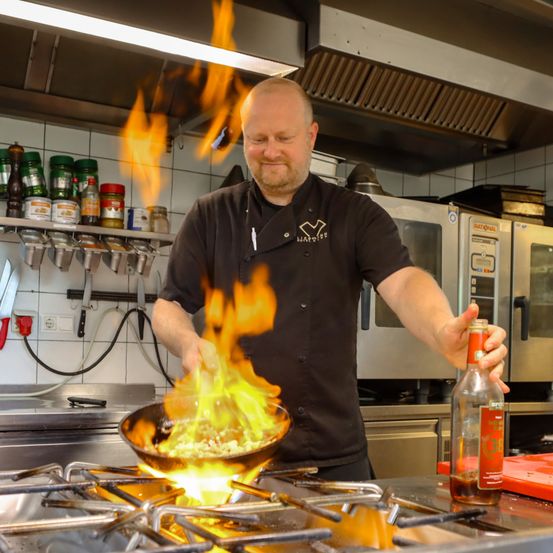 Ein Koch in der Küche, der über einem flammenden Wok steht, fügt seinem Gericht Sauce hinzu, während er ein schwarzes Kochuniform trägt.