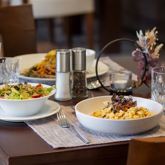 Ein Tisch mit zwei Schüsseln mit Essen, eine mit Salat und eine mit Pasta, umgeben von Gläsern, Salz- und Pfefferstreuer und einer Kerze.