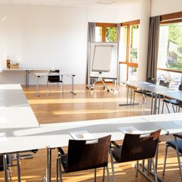 Ein Konferenzraum mit leeren Stühlen, einer Tafel und einem Holzboden. Der Raum ist gut beleuchtet, Sonnenlicht kommt durch die Fenster.