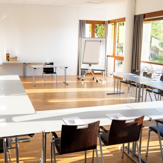 Ein Konferenzraum mit leeren Stühlen, einer Tafel und einem Holzboden. Der Raum ist gut beleuchtet, Sonnenlicht kommt durch die Fenster.