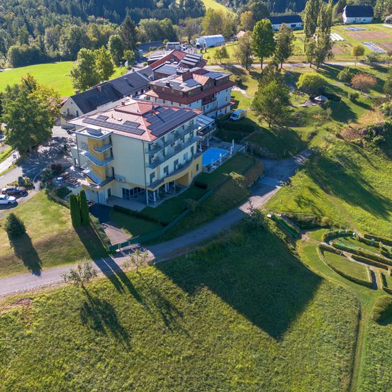 Eine Luftaufnahme eines Hotels mit Swimmingpool und Solarpanels auf dem Dach. Das Hotel ist von einem grünen Feld und Bäumen umgeben. Ein Auto ist an der Seite geparkt.