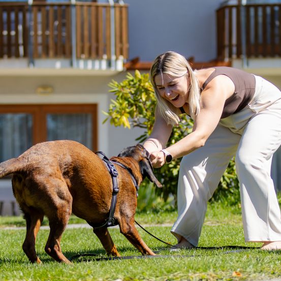 Eine Frau beugt sich vor, um ihren braunen Hund an der Leine in einem Grasbereich zu streicheln.