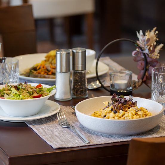 Ein Tisch ist mit verschiedenen Gerichten wie Salat, Nudeln und einem Fleischgericht gedeckt. Salz- und Pfefferstreuer, Gläser und eine Kerze sind ebenfalls vorhanden.