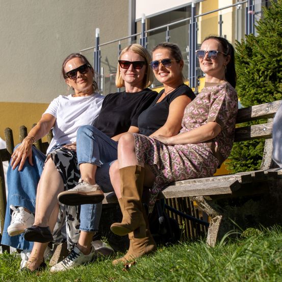 Vier Frauen in Sonnenbrillen sitzen zusammen auf einer Bank im Freien und lächeln für die Kamera. Sie sind von Grün und einem Gebäude mit Glasgeländern umgeben.