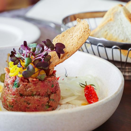 Eine Schüssel mit einem Gericht aus rohem Hackfleisch mit Gemüse, Kräutern und einer Scheibe Brot auf der Seite und einem Korb mit Brot im Hintergrund.