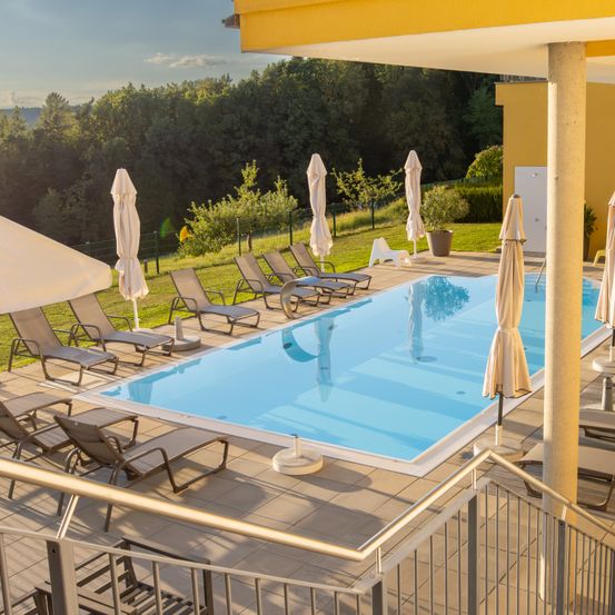 Ein Außenpoolbereich mit Liegestühlen, Sonnenschirmen und einem gelblich gestrichenen Gebäude. Der Pool ist sauber mit blauem Wasser und von grünem Gras umgeben.
