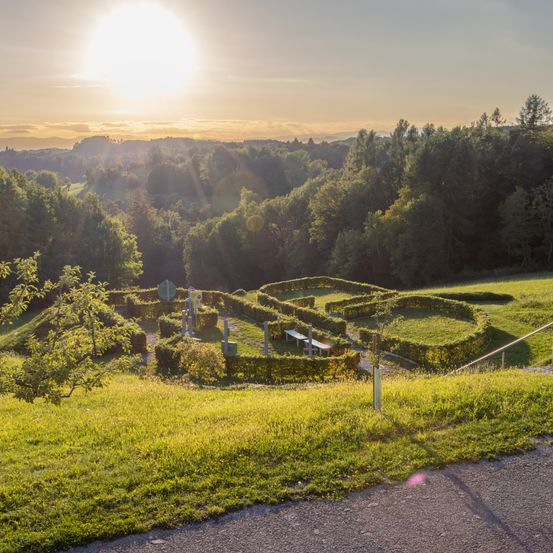 Sonnenuntergang über einer hügeligen Landschaft mit einem großen Labyrinth, umgeben von viel Grün. Eine Bank ist im Labyrinth aufgestellt.