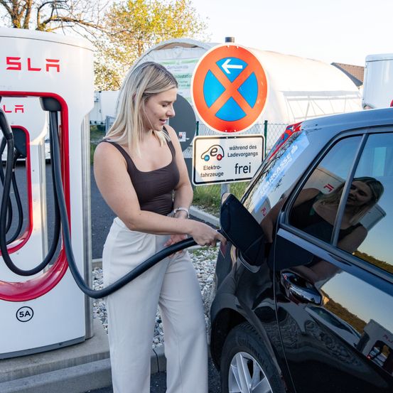 Eine Frau lädt ein Elektroauto an einer Tesla-Ladestation auf. Das Schild zeigt an, dass das Laden während des Be- und Entladens kostenlos ist.