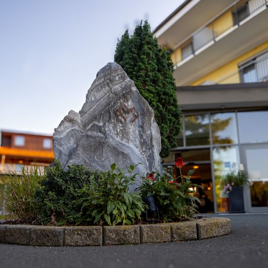 Ein großer Stein mit eingraviertem Text steht in einem Garten mit Blumen und Büschen. Er befindet sich vor einem gelben Gebäude mit Glaswänden.