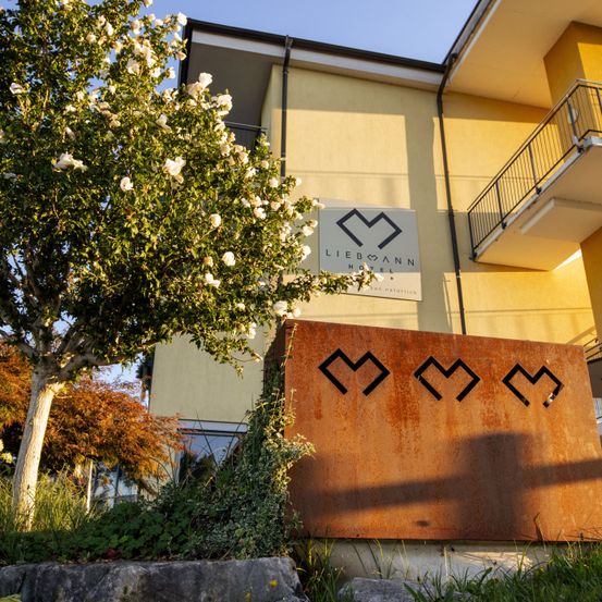 Die Vorderseite des Liebmann Hotels mit einem Schild mit einem abstrakten Design, umgeben von einem Baum mit weißen Blüten und einem Balkon am Gebäude.
