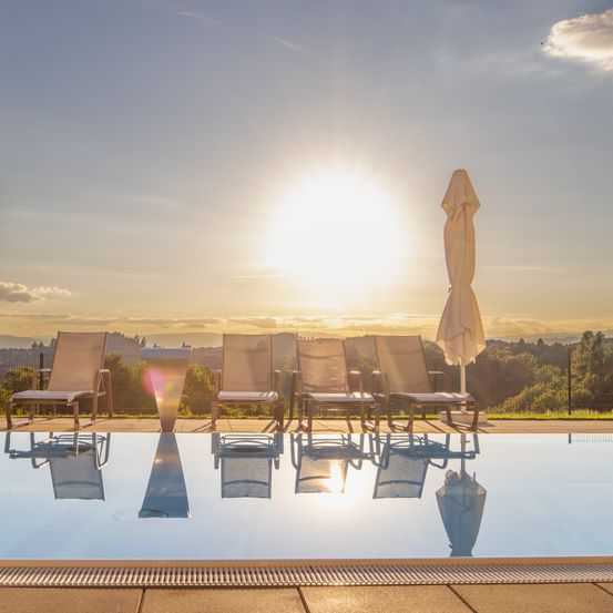 Ein ruhiger Außenpoolbereich bei Sonnenuntergang. Vier Liegestühle stehen am Rand, einer hat einen Schirm. Die Sonne spiegelt sich im Wasser, und der Himmel ist teilweise bewölkt.