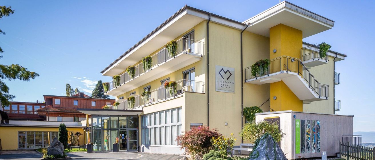 Das Liegmann Hotel, ein gelbes und weißes Gebäude mit Balkonen, befindet sich in einer malerischen Gegend mit Bäumen und Bergen in der Ferne.