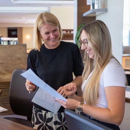 Zwei Frauen in einer Hotellobby, eine hält einen Papierplan und lächelt, die andere trägt eine Brille und lächelt ebenfalls, während sie das Papier hält.