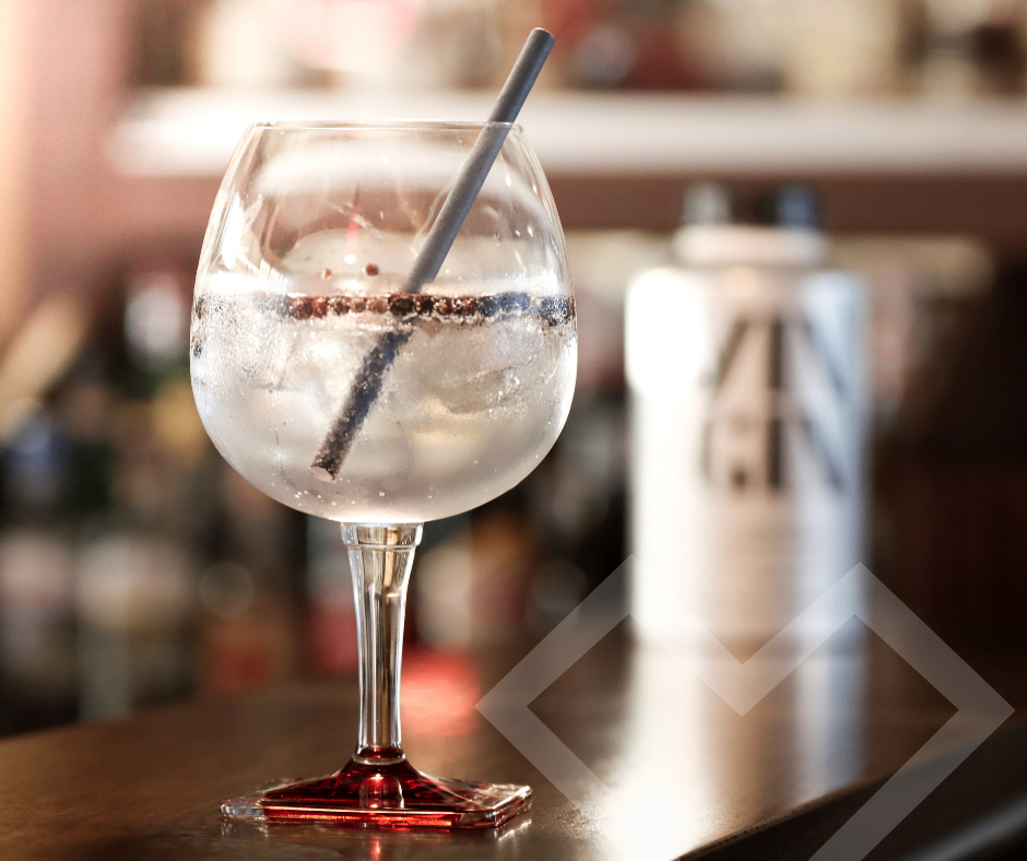 Ein Glas Sprudelwasser mit einem Strohhalm steht auf einer Theke. Im Hintergrund sind verschwommen Flaschen und ein Logo zu sehen.