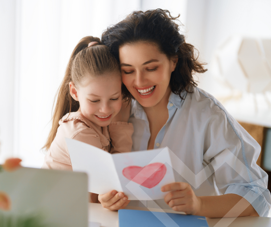 A smiling mother and daughter look at a card with a heart on it. The mother is holding the card, and the daughter is smiling at the camera.