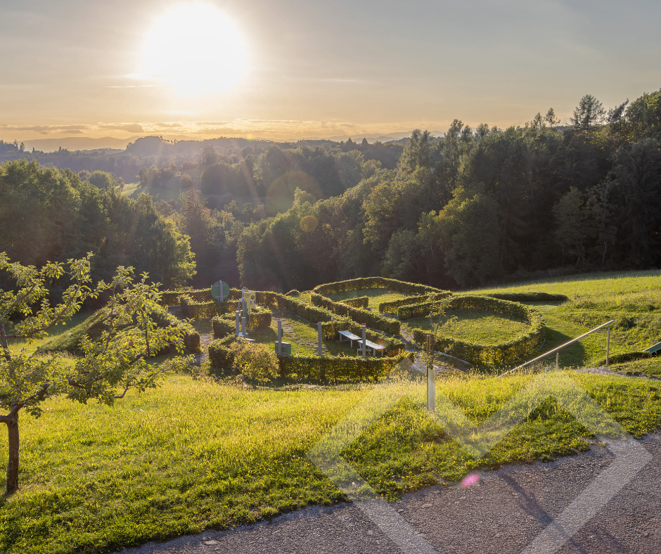 Sonnenuntergang über einem grünen Feld mit einem Labyrinth, Bänken und Bäumen. Eine strahlende Sonne scheint durch die Wolken.