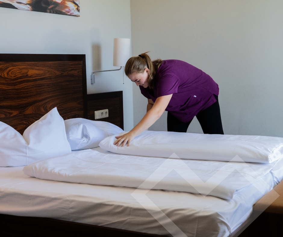 Eine Frau in einem lila Shirt macht ein Bett in einem Hotelzimmer zu. Das Bett hat weiße Laken und Kissen, und das Zimmer hat einen Holzkopfteil und eine weiße Wand.