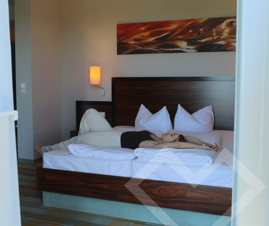Eine Frau liegt auf einem Bett mit weißen Kissen und einer warmen Lampe an der Wand. Das Bett hat einen Holzrahmen und einen karierten Boden.