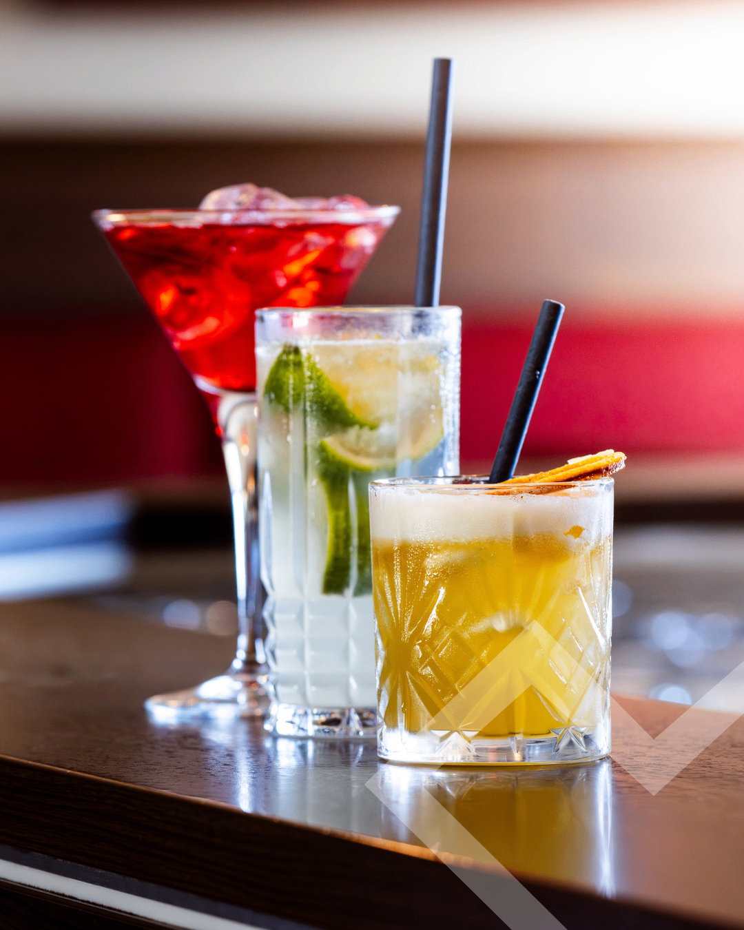 Three cocktails on a bar: a red cocktail in a martini glass, a yellow cocktail with lime slices in a tall glass, and a yellow cocktail with an orange peel in a textured glass.