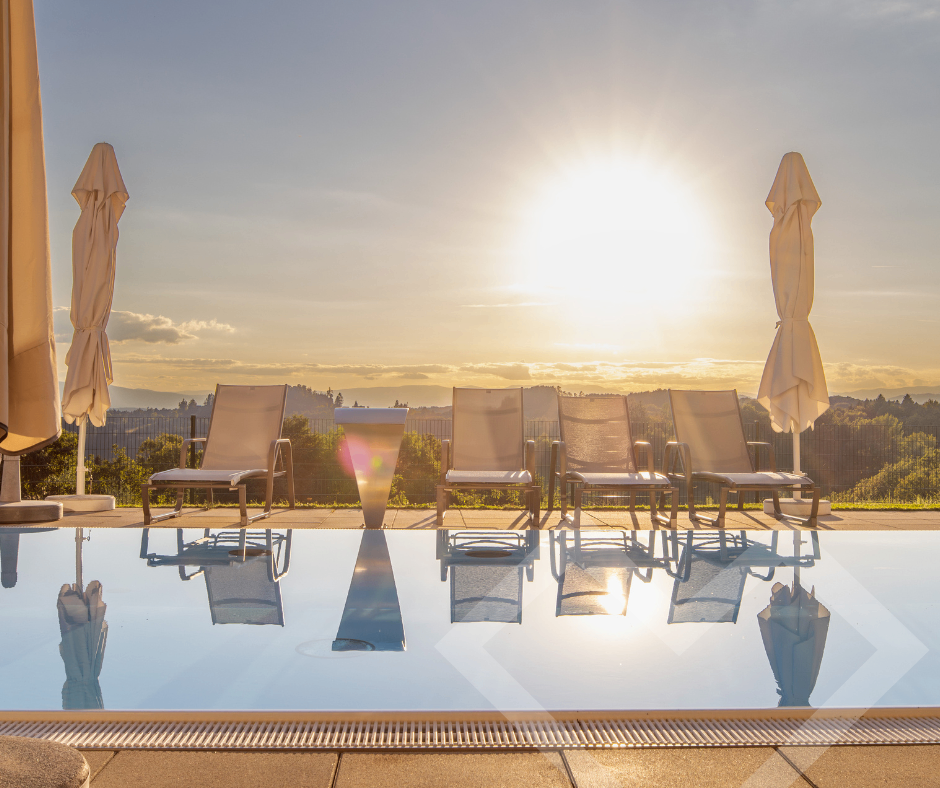 Six lounge chairs and umbrellas line a pool, reflecting in its clear water. The sun sets in the distance over a hilly landscape.