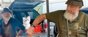 Ein älterer Mann kocht auf einem Grill im Freien mit einem großen Topf. Ein weiterer Mann in einem Hut steht in der Nähe, und ein Lieferwagen ist im Hintergrund.