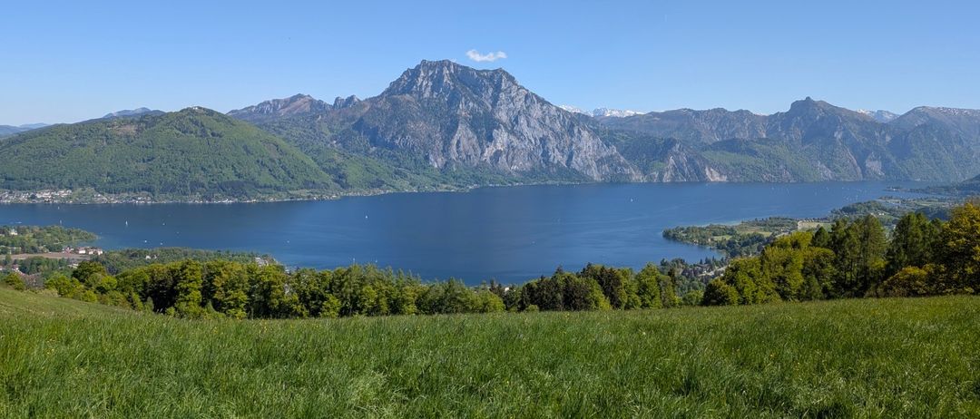 Ein malerisches Panorama eines ruhigen Sees, umgeben von üppigen grünen Hügeln und majestätischen Bergen unter einem klaren blauen Himmel.