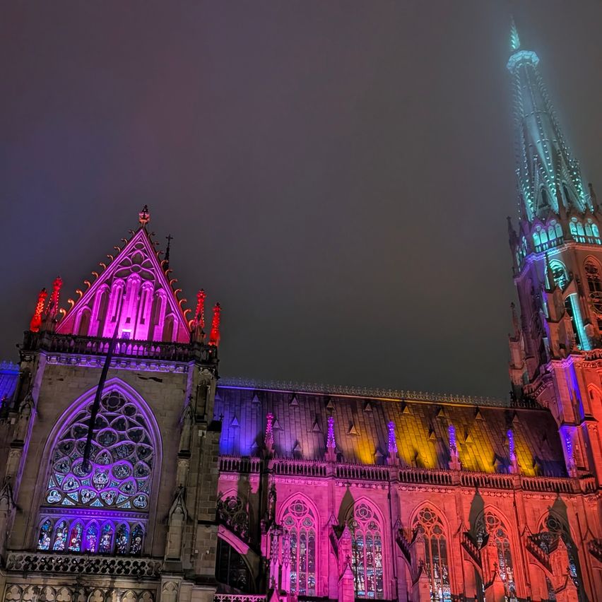 Ein Dom, der bei Nacht mit bunten Lichtern beleuchtet ist. Die Fassade zeichnet sich durch kunstvolle Designs und Spitzbögen aus.