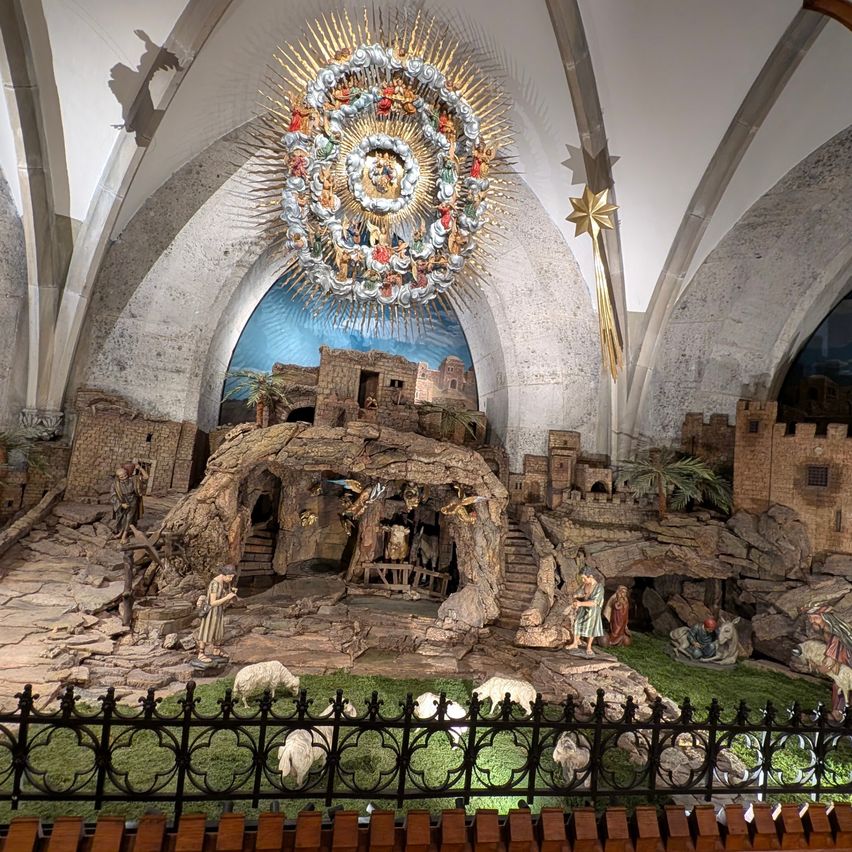 Eine Indoor-Krippenszene mit Figuren in einer Kirche, unter einer goldenen kreisförmigen Dekoration. Die Szene umfasst eine Krippe, Schafe und Menschen. Die Umgebung ist von einem schwarzen Zaun umschlossen.