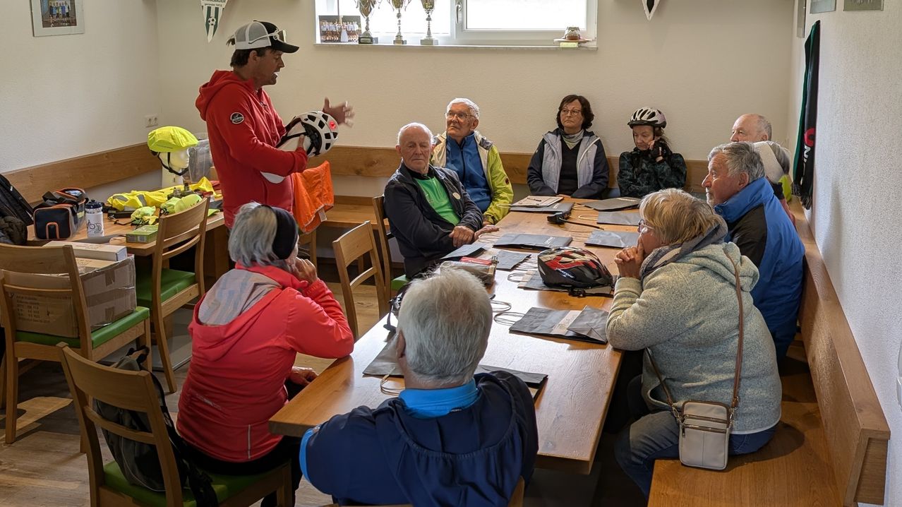 Eine Gruppe von Menschen sitzt um einen Tisch in einem Raum. Ein Mann in Rot hält einen Helm und spricht. Trophäen und Papiere sind auf dem Tisch. Einige tragen Brillen und Helme.