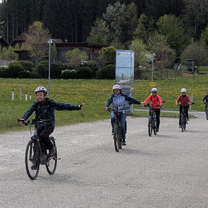 Eine Gruppe von Radfahrern mit Helmen fährt auf einem Kiesweg, umgeben von üppiger grüner Landschaft und Häusern. Der Weg ist gesäumt von Bäumen und Büschen.