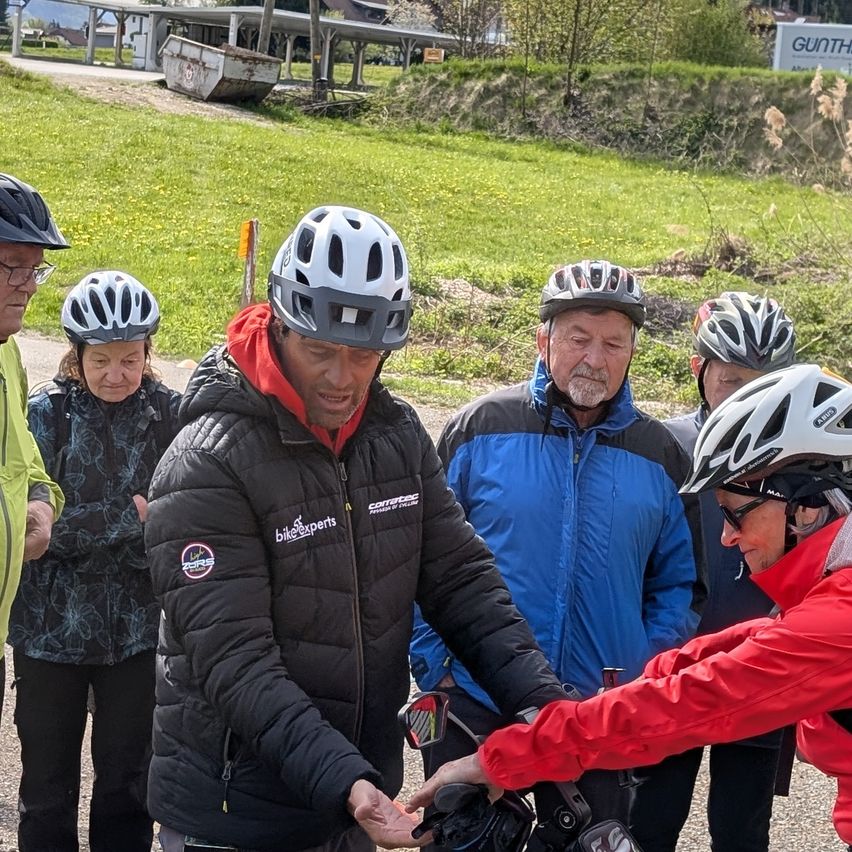 Eine Gruppe von Personen mit Helmen ist auf einer Straße versammelt, zwei halten ein Fahrrad. Eine Person trägt eine rote Jacke, und eine andere hat eine schwarze Jacke mit sichtbarem Logo.