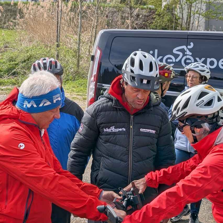 Fünf Personen in roten Jacken stehen um einen schwarzen Van mit weißem Logo und unterhalten sich. Zwei von ihnen tragen Helme und Schutzbrillen. Der Mann in der Mitte hält ein Gerät. Im Hintergrund sind Bäume und Büsche zu sehen.